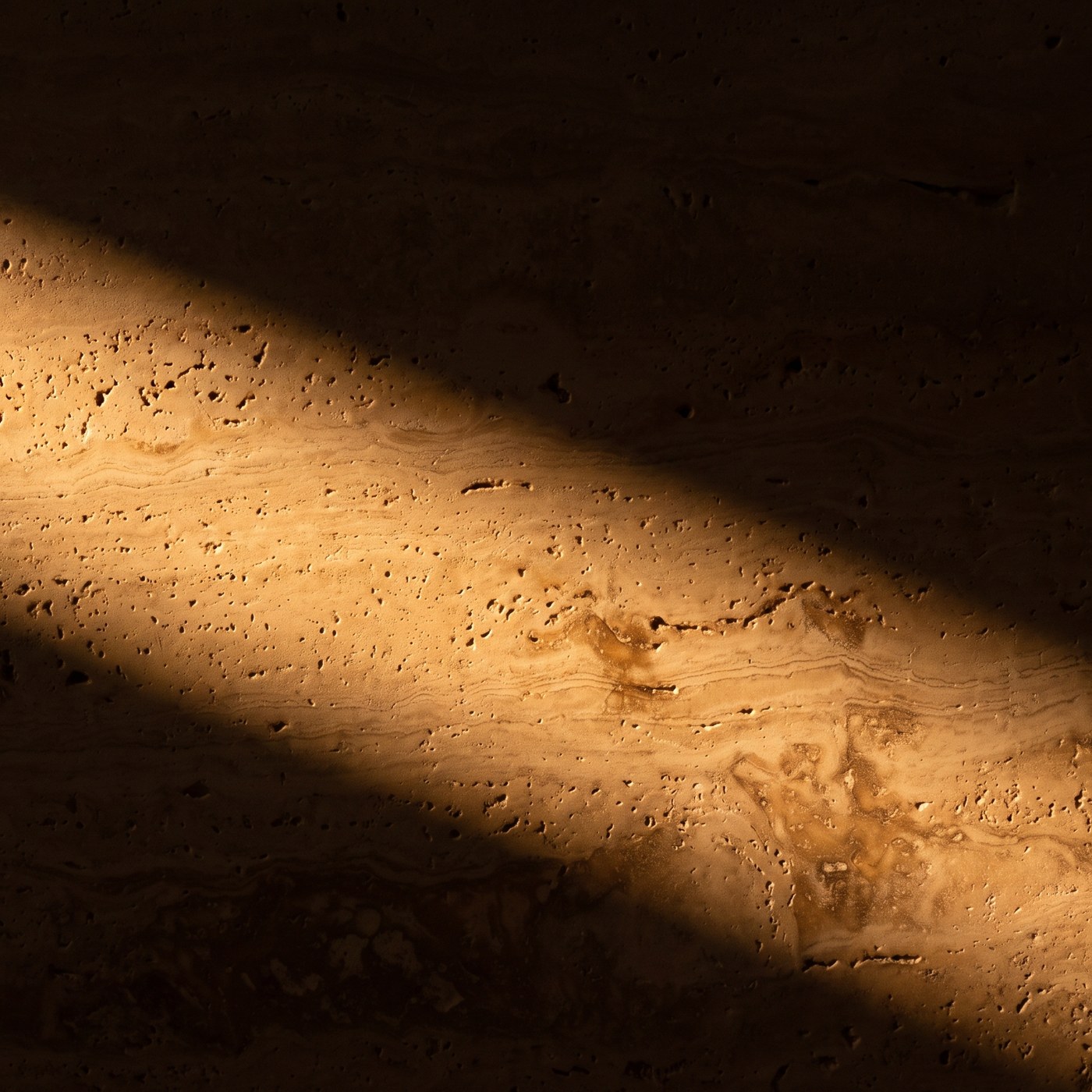 Close detail of honed travertine, its open pores catching low warm light.