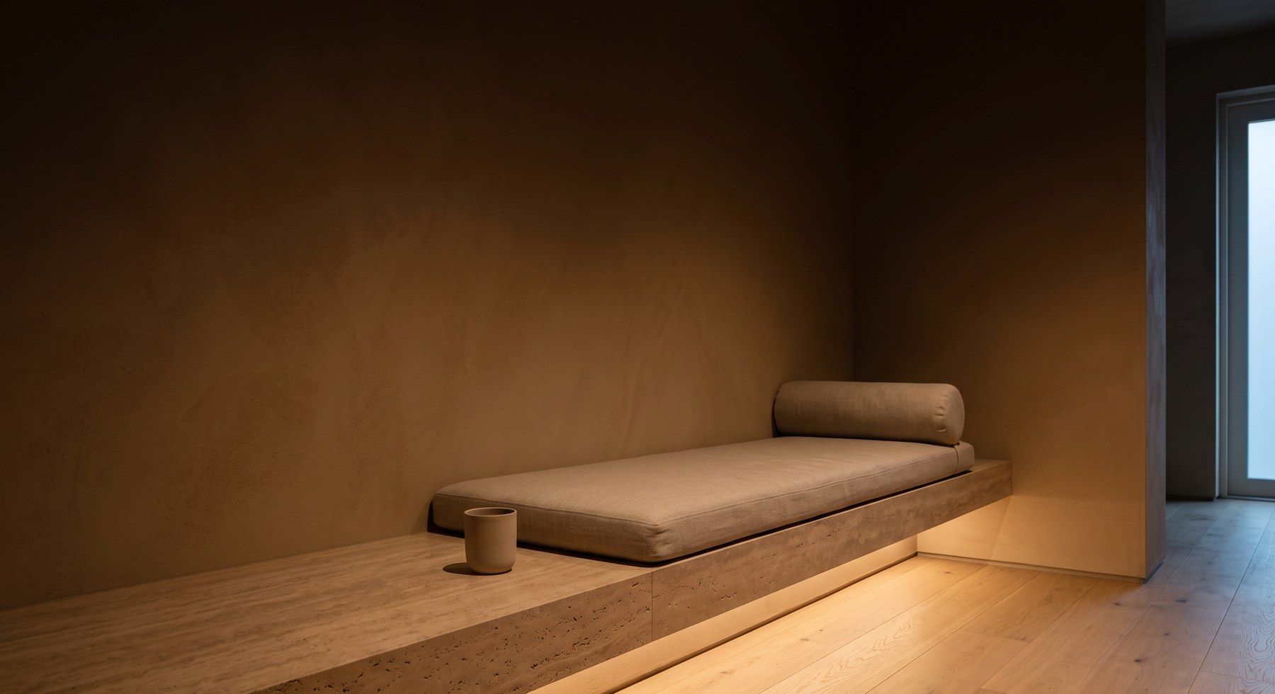 The warm room at Northlight: a travertine bench with a linen cushion and a ceramic cup, lit by a single strip of light near the floor.