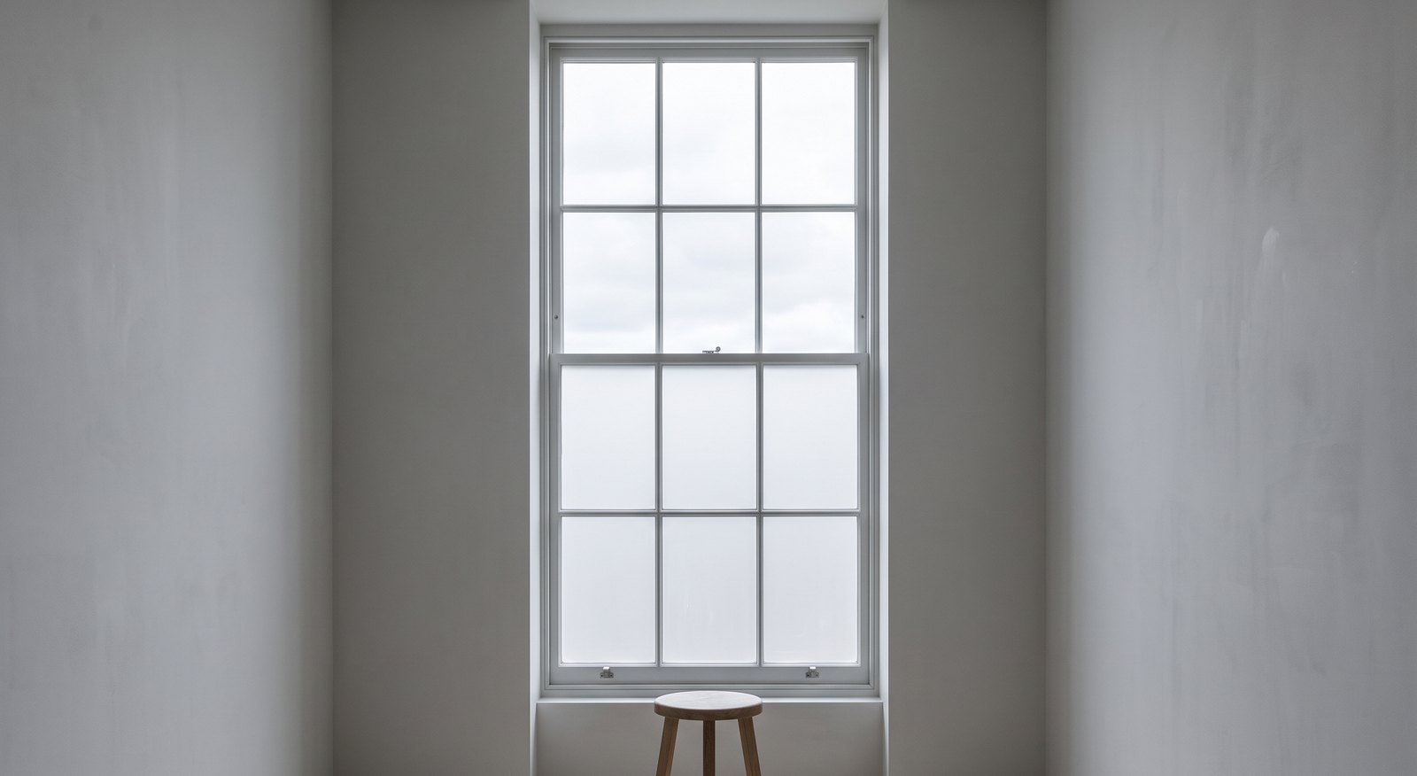 The north room at Northlight, empty: a tall sash window with frosted glass, plain grey plaster walls and a single wooden stool on the floor.
