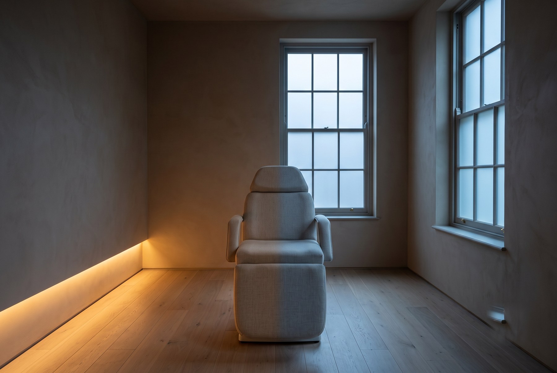 The north room at Northlight: a plain treatment chair on oak boards between two tall sash windows, with one warm strip of light low on the left wall.